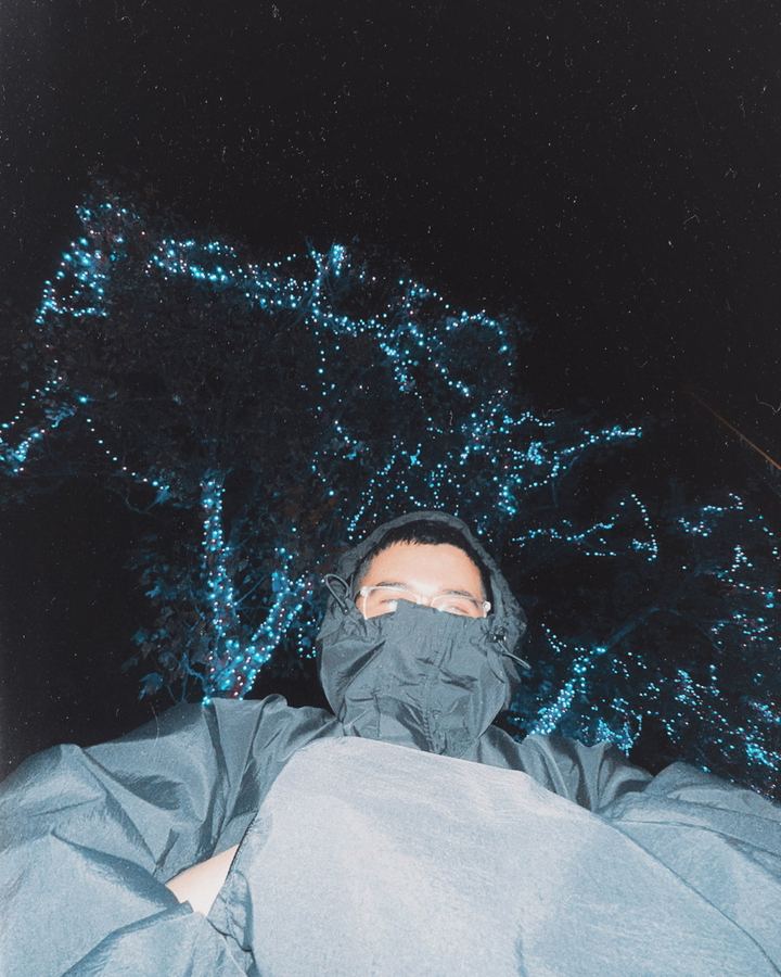 A hooded figure in glasses below a tree wrapped in blue lights.