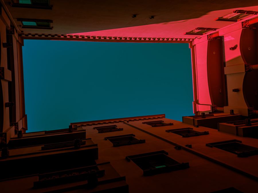 Looking straight up from a courtyard at a teal sky framed by red-lit walls.