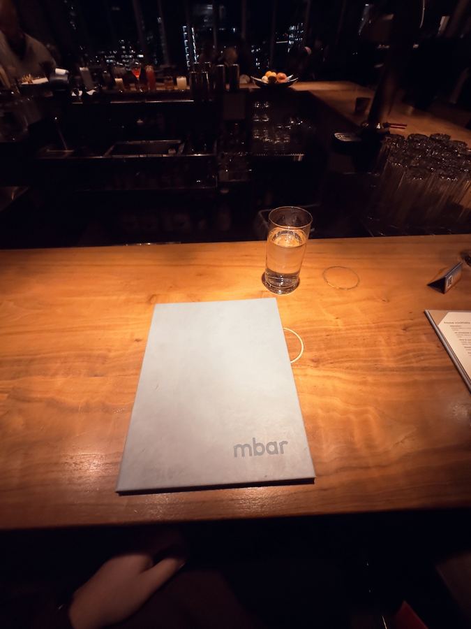 A pale blue menu on a warm-lit wooden bar beside a glass of water.