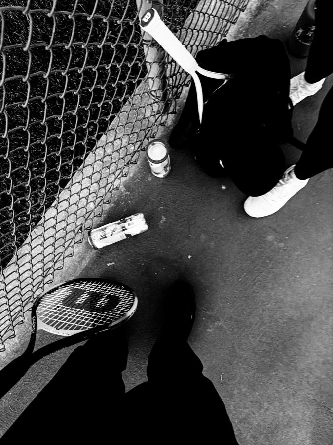 Rackets, ball cans and sneakers beside a chain-link fence in black and white.