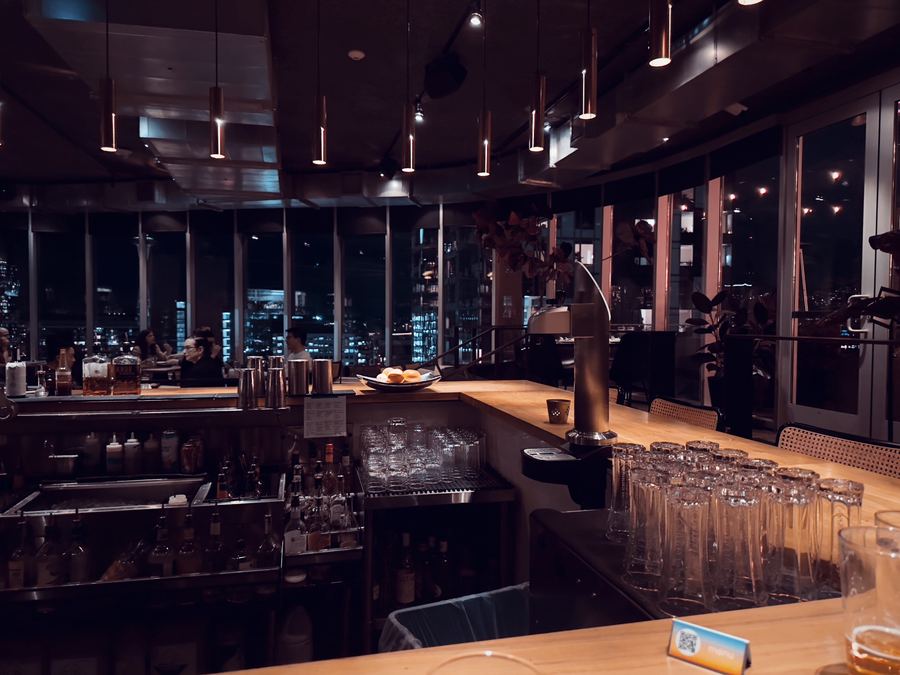 A bar with copper pendant lights and stacked glasses, city lights through the windows.