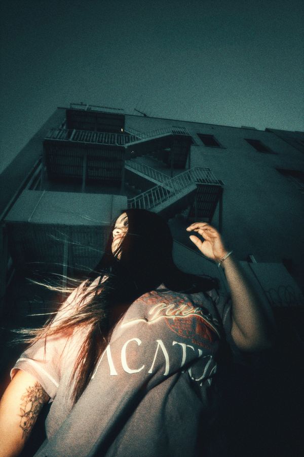 Hair flying, one hand raised, beneath zigzag fire escape stairs.